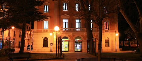 Un edificio histórico con una entrada central, un patio con bancos y una fachada bien iluminada.