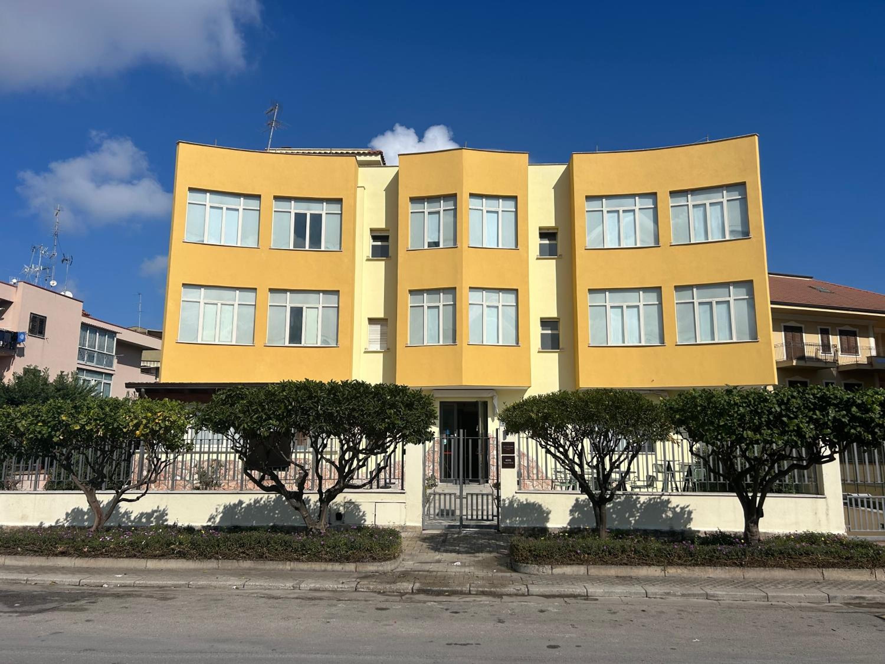 Un edificio amarillo de varios pisos con fachada simétrica, ventanas grandes y entrada protegida por una reja.