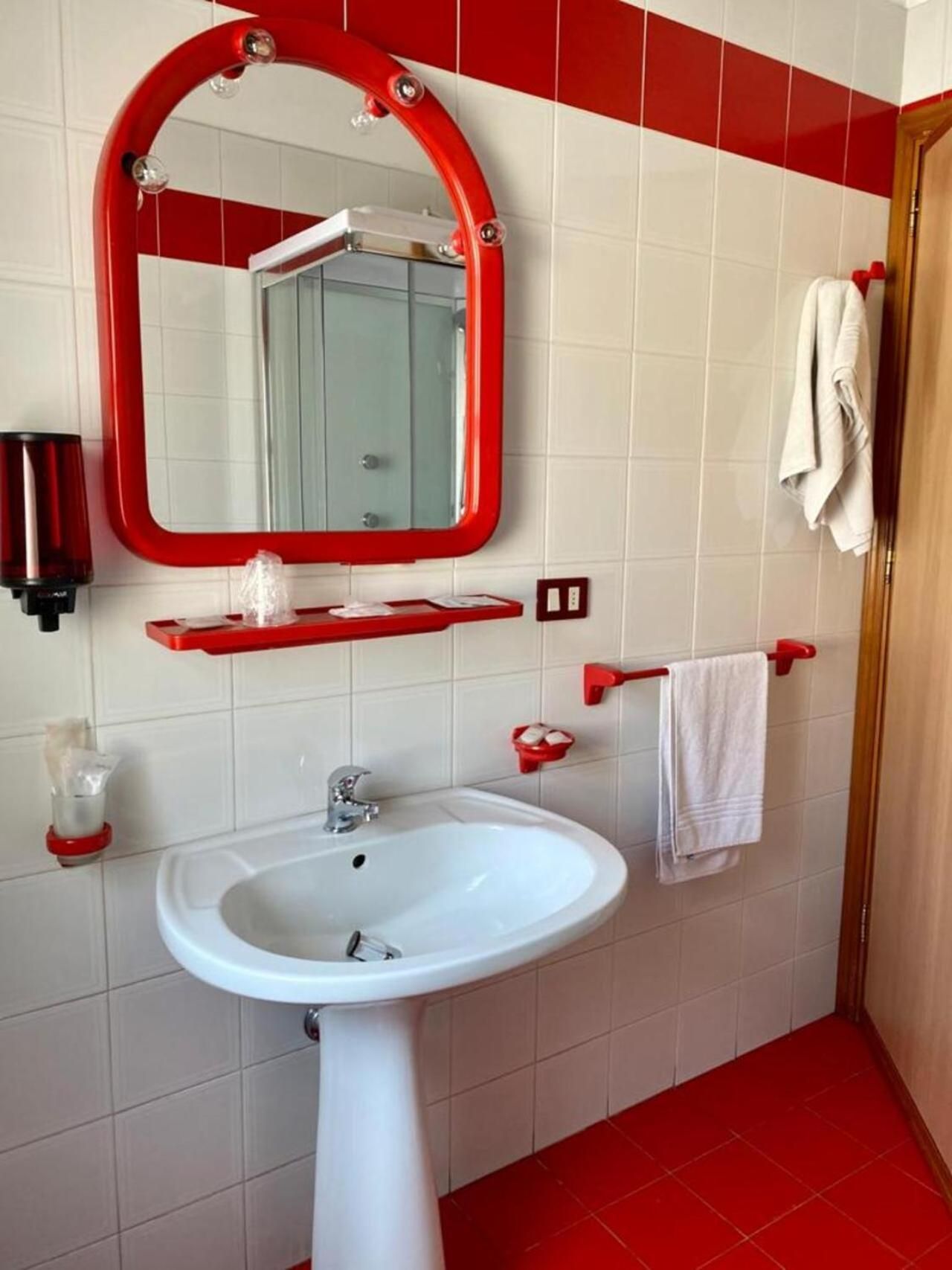 Un bagno con un lavandino bianco, pareti piastrellate di rosso e uno specchio con cornice rossa, oltre a un portascopino.