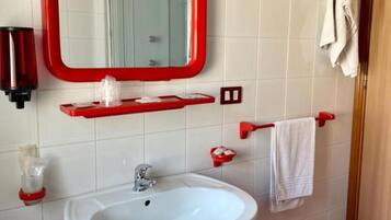 Un bagno con un lavandino bianco, pareti piastrellate di rosso e uno specchio con cornice rossa, oltre a un portascopino.