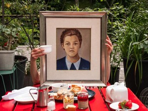 A framed portrait being held up, with a table in front featuring various food items and a teapot. - Frida Am Wald (Rasun Anterselva)