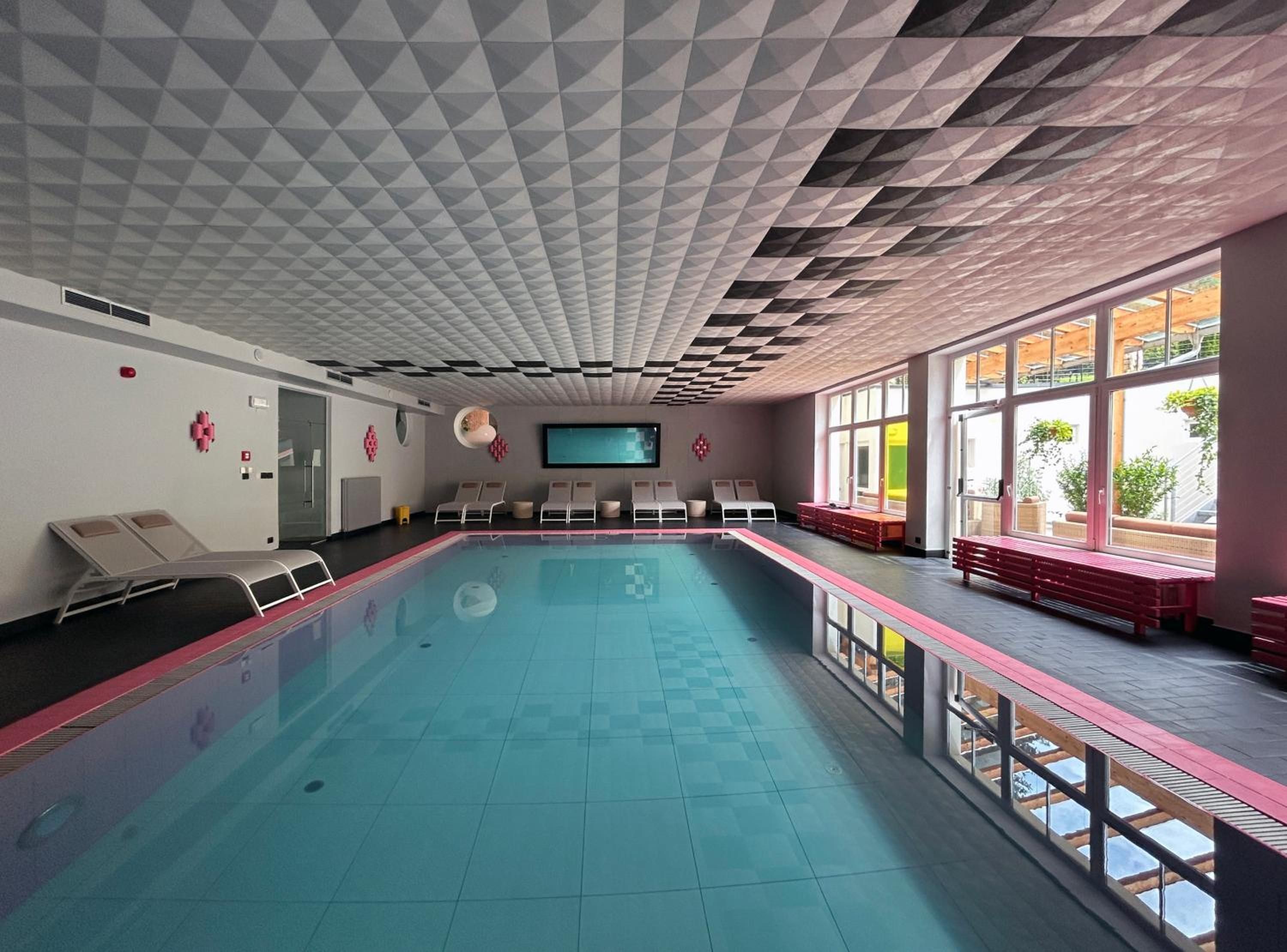 Une piscine bordée de chaises longues et de bancs, de grandes fenêtres et un plafond orné d’un motif géométrique.