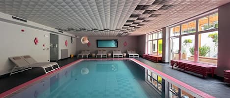 Une piscine bordée de chaises longues et de bancs, de grandes fenêtres et un plafond orné d’un motif géométrique.