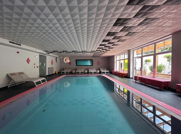 A swimming pool with lounge chairs and benches along the sides, large windows, and a geometric ceiling pattern. - Frida Am Wald (Rasun Anterselva)