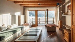 A room with a wooden floor, a bed with pillows, a bench, a table, and a view of a snowy landscape through a large window. - Frida Am Wald (Rasun Anterselva)