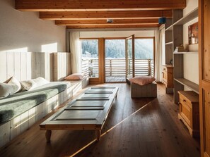Une pièce avec un plancher en bois, un lit avec des oreillers, un banc, une table et une vue sur un paysage enneigé par une grande fenêtre.