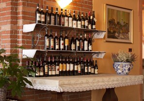 Una estantería para vinos con varias botellas frente a una pared de ladrillo, una mesa con mantel de encaje y una planta en maceta.