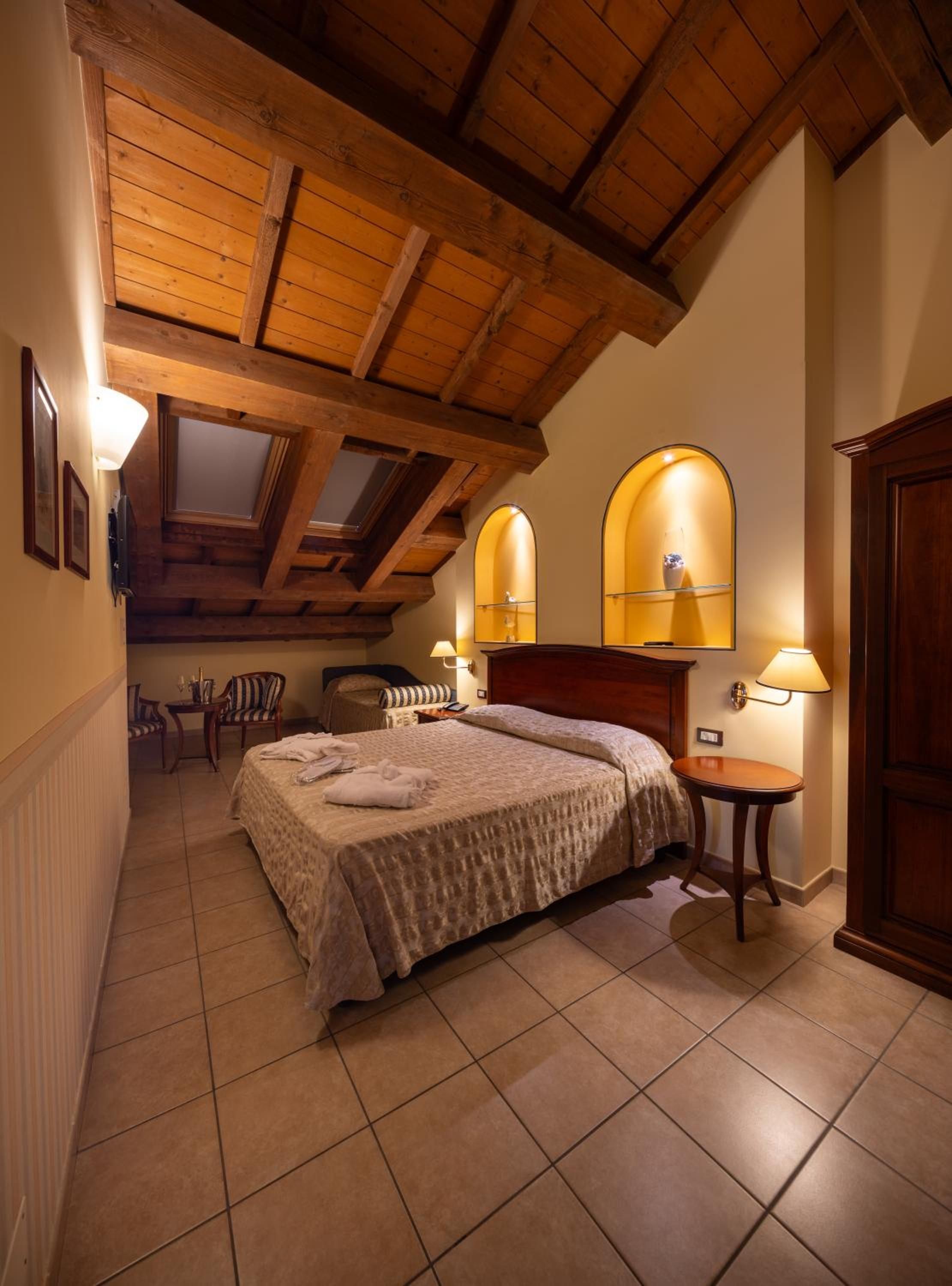 A bedroom with a wooden ceiling, a bed with a headboard, two bedside tables with lamps, a chair, and a small table with a lamp.