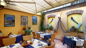 Un área de comedor al aire libre con mesas preparadas para dos, cubiertas con manteles azules y rodeadas de sillas de mimbre.