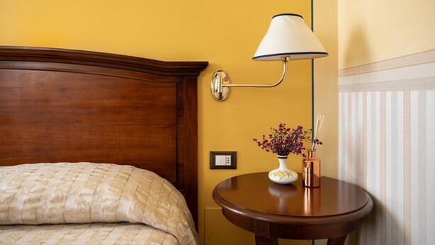 Una habitación de hotel con cabecera de madera, una mesita de noche con un jarrón de flores y una lámpara de pared.