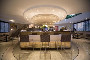 Un moderno vestíbulo de hotel con una zona de bar central, asientos y una escalera que lleva a un nivel superior.