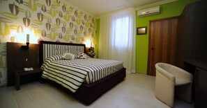 A bedroom with a bed, bedside lamps, a nightstand, a chair, and a wall-mounted TV. - Hotel Tropical (Ostuni)