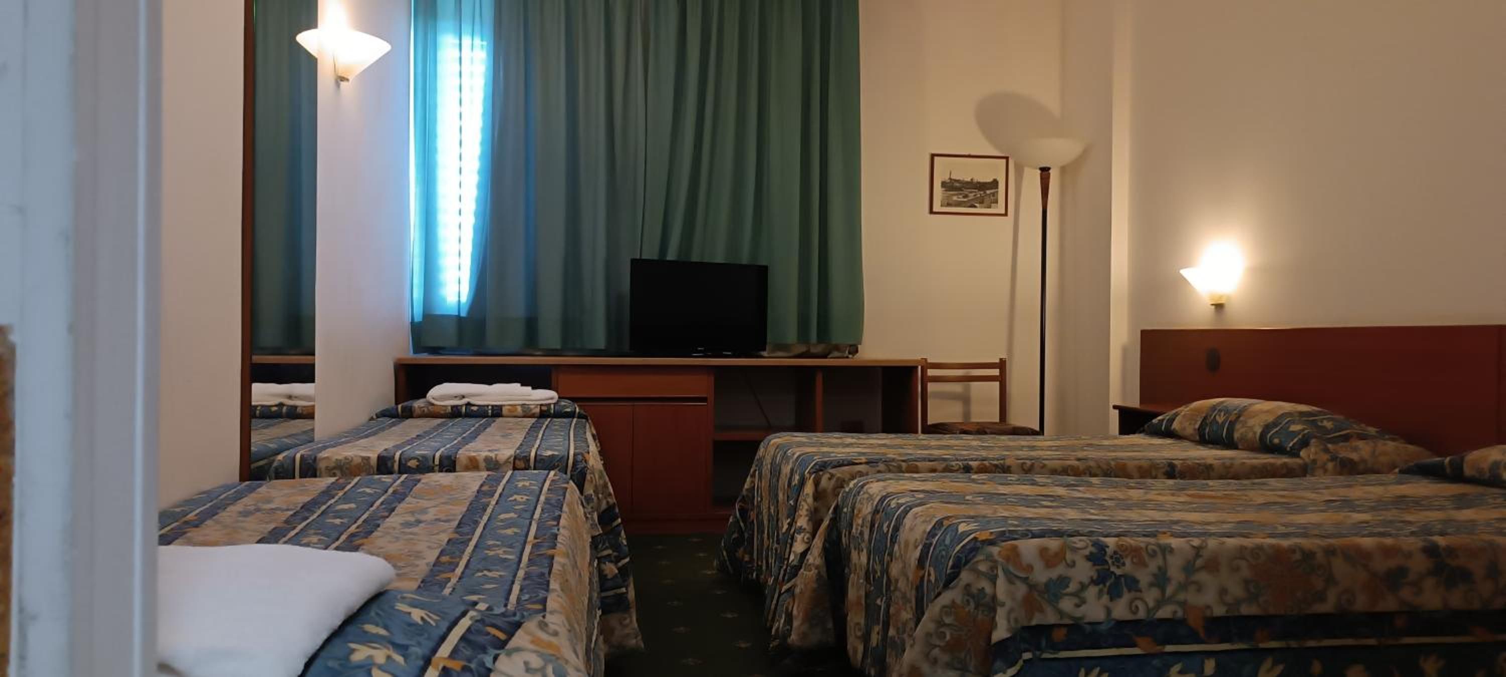 Una camera d'albergo con due letti, una scrivania con televisore e una lampada da terra.