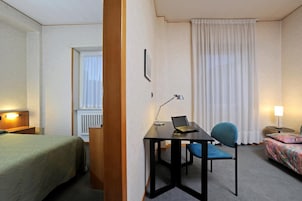 Habitación de hotel con cama, escritorio con laptop, silla y mesita de noche con lámpara.