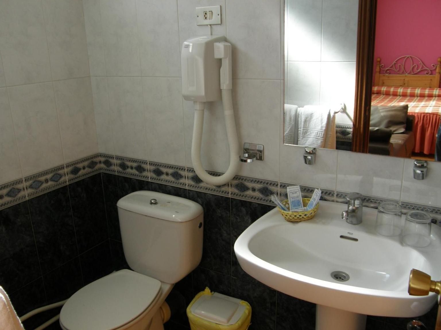 Egy fürdőszoba, amelyben található egy WC, egy mosdókagyló és egy tükör, amelyben egy piros fejrésszel ellátott ágy képe látható.
