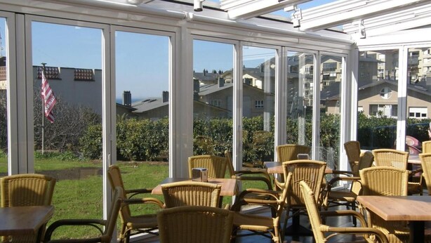 A conservatory with wicker furniture, large windows, and a view of residential buildings. - Hotel Goizalde (Sopelana)