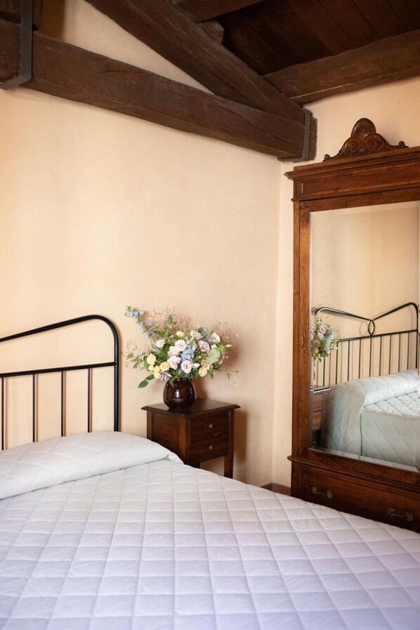 Un dormitorio con cama, mesita de noche de madera, un jarrón con flores y un espejo de cuerpo entero.