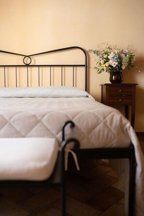 Una cama con colchón acolchado y cabecera de metal, una mesita de noche de madera con un jarrón de flores y suelo de baldosas.