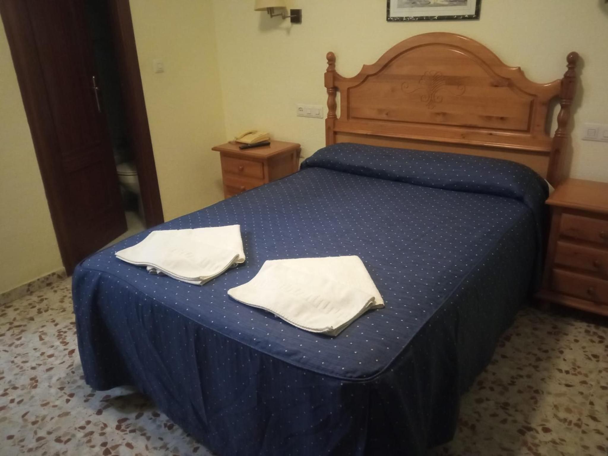 Un dormitorio con cabecera de madera, cama con colcha azul, dos toallas blancas sobre la cama, una mesita de noche de madera con un teléfono, y un pequeño mueble con cajones.