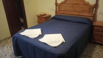 Un dormitorio con cabecera de madera, cama con colcha azul, dos toallas blancas sobre la cama, una mesita de noche de madera con un teléfono, y un pequeño mueble con cajones.