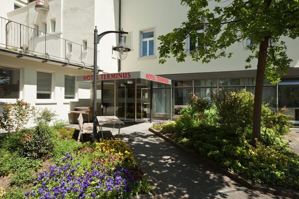 Un hotel llamado 'HOTEL TERMINUS' con una fachada moderna, zona de estar al aire libre y un jardín bien cuidado.