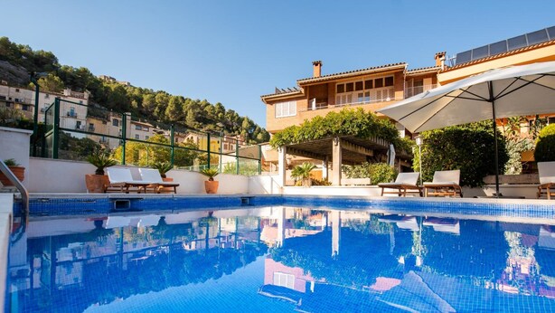 Ein Schwimmbecken mit Liegestühlen, einer Pergola und einem Gebäude mit einem Ziegeldach im Hintergrund.