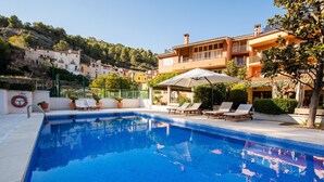 Ein Swimmingpool mit Liegestühlen und Sonnenschirmen, umgeben von Gebäuden und einer Hügelkuppe.