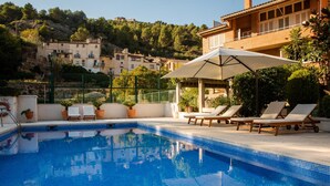 Ein Swimmingpool mit Liegestühlen und einer Pergola, umgeben von Grünflächen und Gebäuden im Hintergrund.