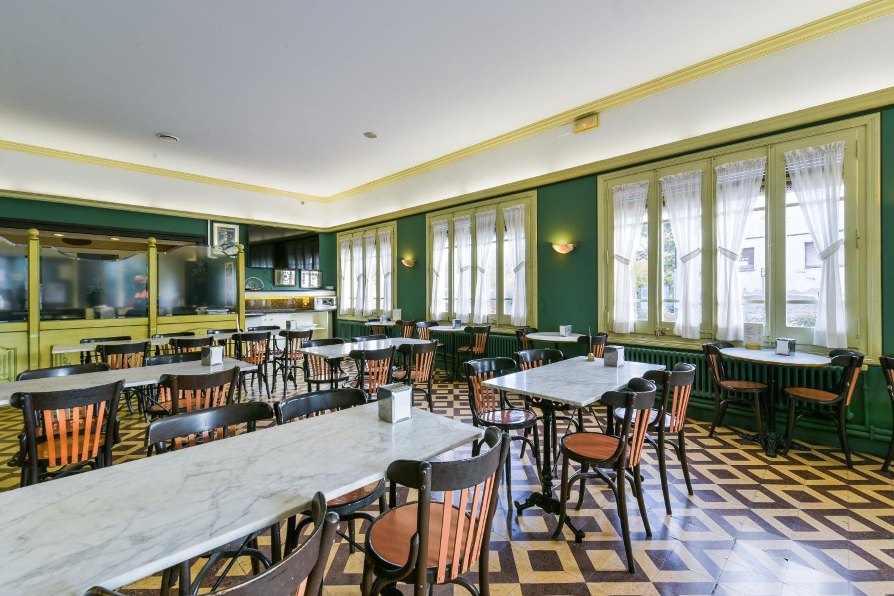A dining area with multiple tables and chairs, large windows with curtains, and a kitchen area visible in the background.