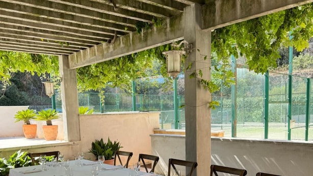 A long dining table set for a meal, surrounded by wooden chairs, under a covered area with a view of greenery and a fence. - Hotel Robert (La Pobla de Claramunt)