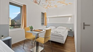 Habitación de hotel con cama, un escritorio y dos sillas, una mesita con una planta, y vista a la montaña a través de la ventana.