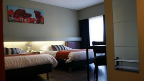 A hotel room with two beds, a desk, and a chair. There is a painting on the wall and a window with curtains. - De Basiliek (Edegem)