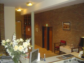 A reception area with a brick wall, a reception desk, a chair, a table with a lamp, and a framed picture on the wall. - De Basiliek (Edegem)