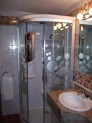 Un baño con ducha envuelta en vidrio, un lavamanos con encimera de granito y un espejo con detalles de azulejos florales.
