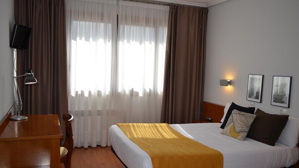 A hotel room with a bed, a desk, a chair, a lamp, a window with curtains, and wall-mounted lights. - Hotel Elorrio (Elorrio)