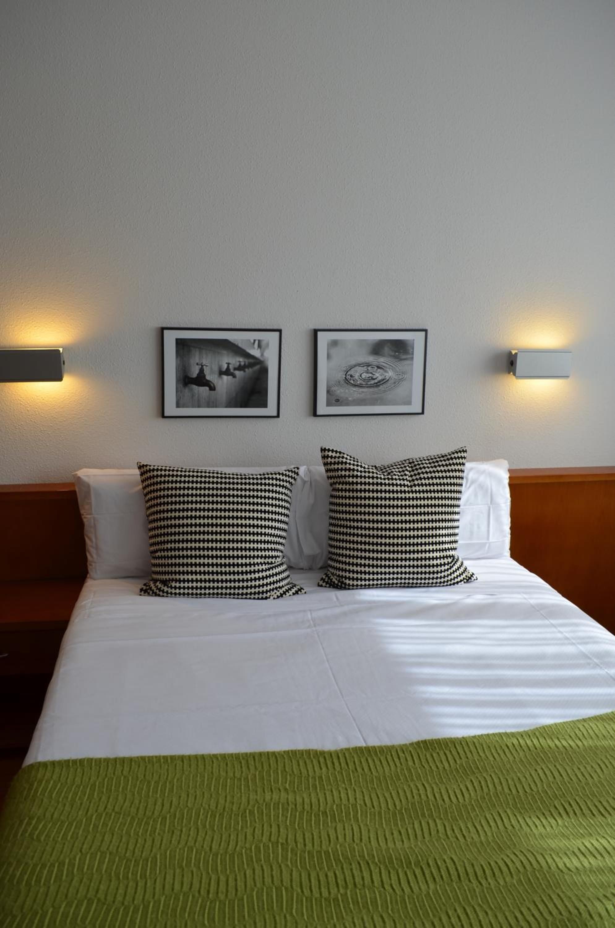 Habitació d'hotel amb un llit, dues almoines, dues làmpades penjades a la paret i dues imatges amb marc.