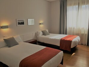 Habitación de hotel con dos camas, una mesita de noche, una lámpara de pared y un ventanal con cortinas.