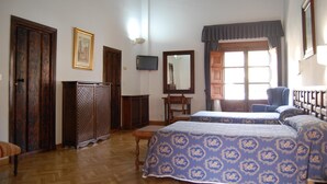 A room with two beds, a wooden wardrobe, a television, and a window with curtains. - Hospederia Del Real Monasterio (Guadalupe)