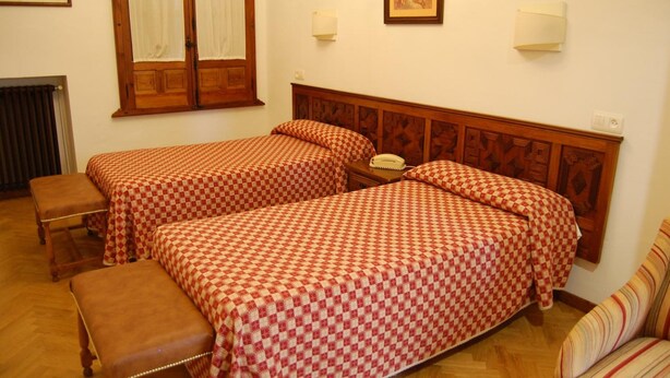 A room with two beds, a bench, a telephone, and a radiator. - Hospederia Del Real Monasterio (Guadalupe)