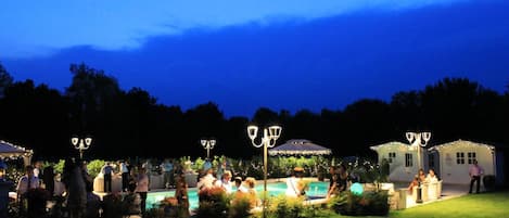 An outdoor gathering with tables and chairs, a pool, and decorative lighting.
