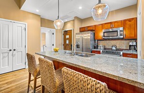 A modern kitchen with a granite countertop, stainless steel appliances, and a woven chair. - Maui Resort Rentals: Expansive 5BR Ground-Floor Retreat with Three Lawn Areas, Steps from Beach and Pools! (Lahaina)