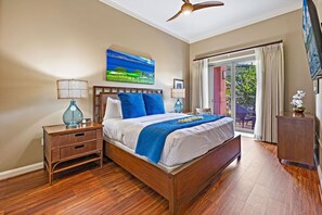 Ein Schlafzimmer mit Bett, Nachttischen, Deckenventilator, einem Gemälde, einem Fenster mit Vorhängen und Blick auf Grünfläche.