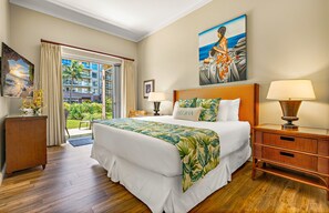 A hotel room with a large bed, a nightstand, a lamp, a painting, and a view of a building and greenery outside. - Maui Resort Rentals: Expansive 6BR Ground-Floor Retreat with Huge Lawn Areas, Steps from Beach and Pools! (Lahaina)