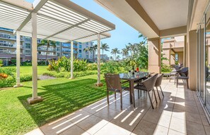 An outdoor patio area with a table and chairs, surrounded by a well-maintained lawn and tropical plants. - Maui Resort Rentals: Expansive 6BR Ground-Floor Retreat with Huge Lawn Areas, Steps from Beach and Pools! (Lahaina)