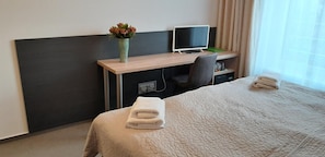 Et hotelværelse med en seng, et skrivebord med en computer, en stol, en vase med blomster og et vindue med gardiner.