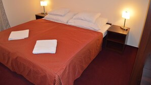 A neatly made bed with a red patterned bedspread, two white pillows, and two white towels folded on the bed. There are two bedside tables with lamps on either side of the bed. - Hotel Aréna (Prague)