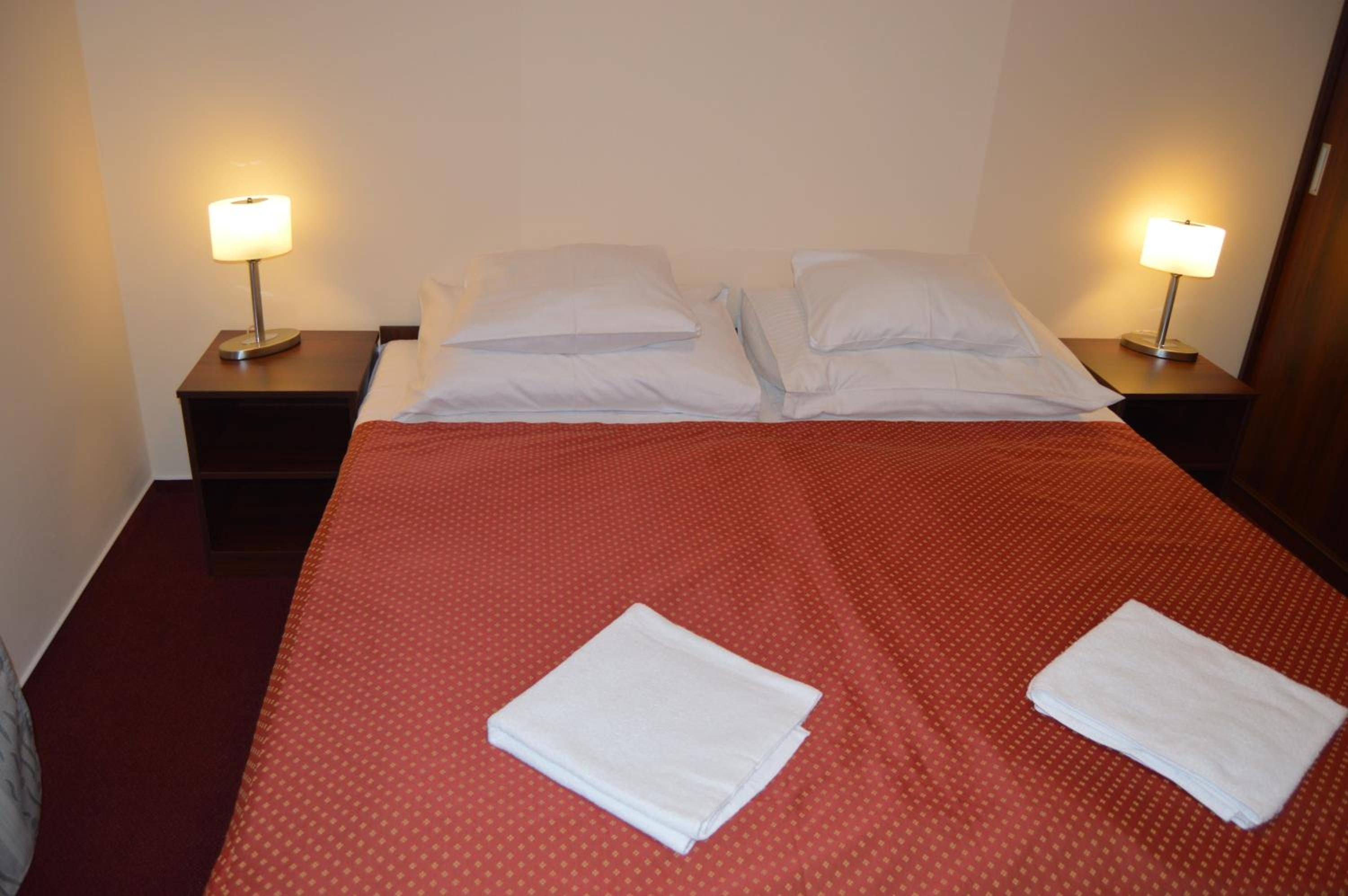 Ein Hotelzimmer mit einem ordentlich bezogenem Bett, zwei Kissen, zwei Nachttischen mit Lampen und zwei gefalteten Handtüchern auf dem Bett.