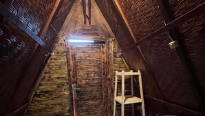 A small, cozy attic bedroom with a wooden sloped ceiling, a single bed, a ladder, and a door leading to a staircase.