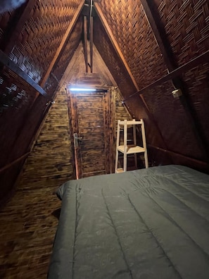 A small, cozy attic bedroom with a wooden sloped ceiling, a single bed, a ladder, and a door leading to a staircase.