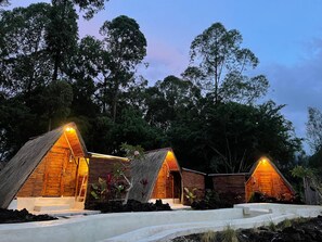 A bungalow-style accommodation with a thatched roof, surrounded by lush greenery and a clear sky.